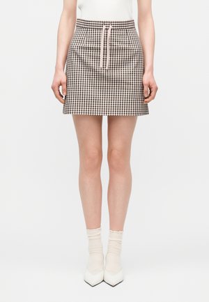 Ženska, oblečena v mini krilo z rjavo-belim gingham vzorcem in roza vrvico, kombinirano z belimi gležnarji in ostrimi belimi čevlji.