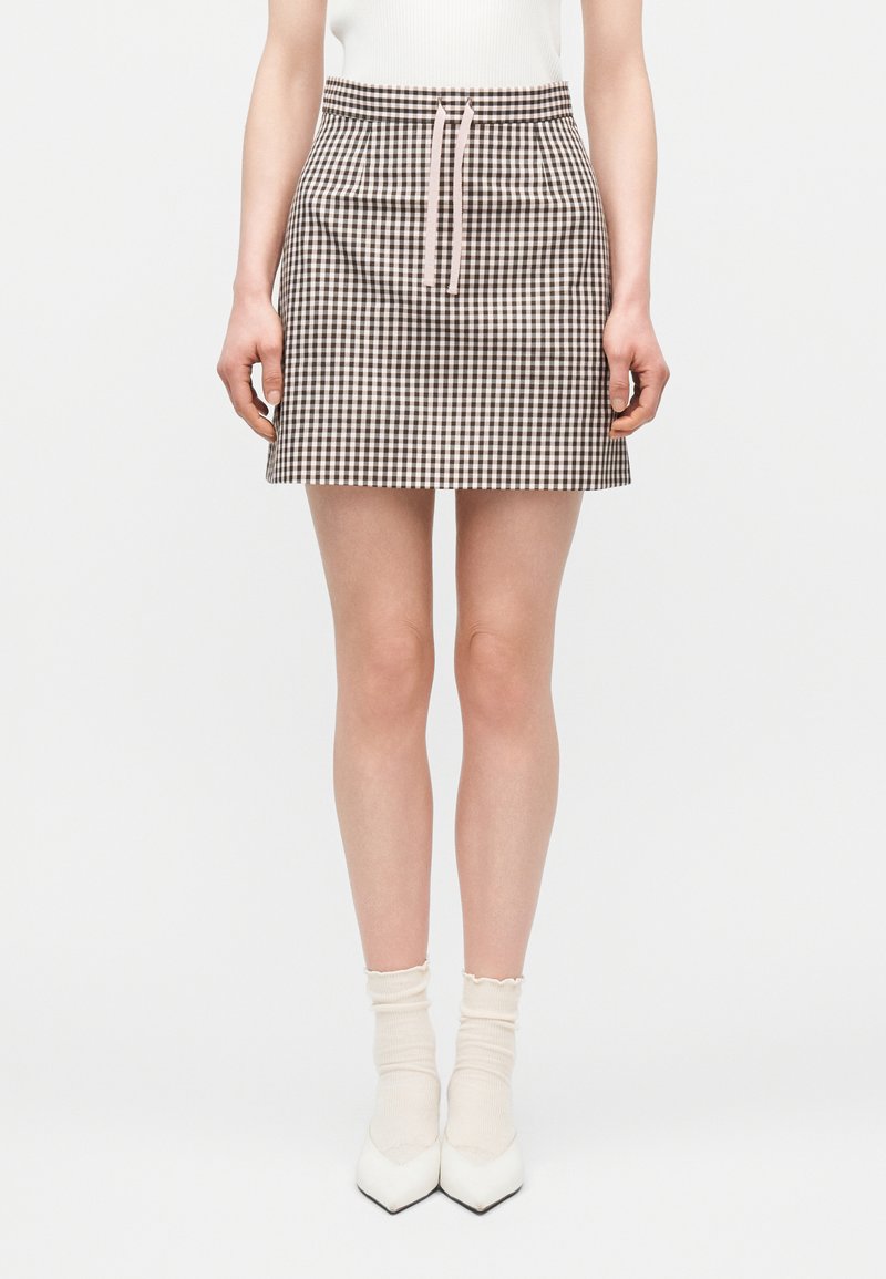 Γυναίκα που φορά μια κοντή φούστα gingham σε καφέ και άσπρο χρώμα με ροζ κορδόνι, συνδυασμένη με λευκές κάλτσες αστραγάλου και μυτερά λευκά παπούτσια.