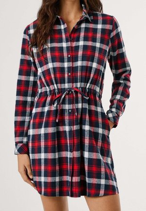 Kobieta ubrana w czerwoną, granatową i białą koszulową sukienkę w kratę z długim rękawem, paskiem w talii i kieszeniami, stojąca na jednolitym tle.