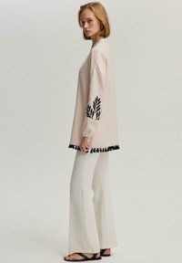 Top oversize crema con ricami a foglie nere e rifiniture con frange nere, abbinato a pantaloni bianchi a zampa d'elefante e sandali neri.