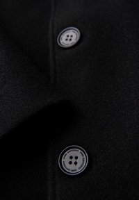 Zwei schwarze Mantelknöpfe mit weißen Rändern und dem Text "OMBRE QUINTESSENCE" auf schwarzem Wollstoff.