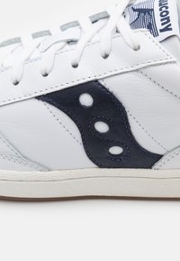 Baskets en cuir blanc avec accent latéral en daim bleu marine, présentant trois découpes rondes, lacets plats blancs, semelle en caoutchouc texturée et détails cousus.