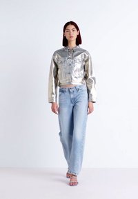 Silver metallic kort jacka med knappstängning, parat med ljusblå denimjeans. Modellen bär sandaler med genomskinliga remmar, minimal bakgrund.