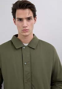 Olivengrøn, knap-op jakke med rundet krave, lavet af glat stof. Har lyse knapper og et enkelt, rent design.