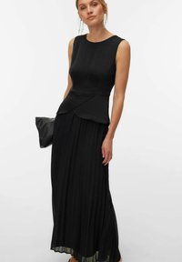 Femme portant un haut noir sans manches à basque et une longue jupe plissée, tenant une pochette noire sur un fond blanc uni.