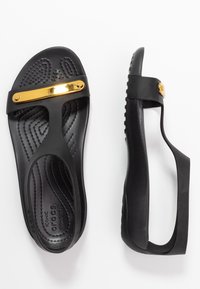 Mustat kumiset sandaalit kultaisella metallisella yksityiskohdalla, joissa on tekstitettujen pohja, minimalistinen muotoilu ja T-nauha tyyli.