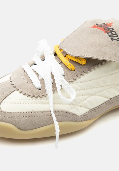 Baskets beige et blanc cassé avec des lacets blancs épais, des lacets jaunes en dessous, et une languette en suède à logo.