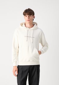 Sudadera con capucha color crema, con un bolsillo frontal y un logo bordado. Material de mezcla de algodón, corte relajado, puños y dobladillo acanalados.