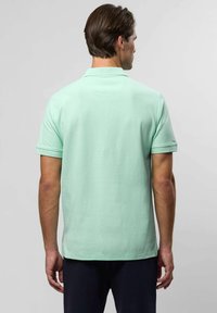 Mężczyzna widziany z tyłu, ubrany w jasnoszary polo z krótkim rękawem i ciemne spodnie, na jednolitym jasnym tle.