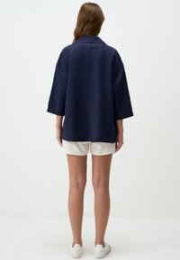 Giacca camicia in lino blu navy con maniche a tre quarti, vestibilità rilassata e collo alla francese, abbinata a pantaloni corti bianchi e scarpe da ginnastica.