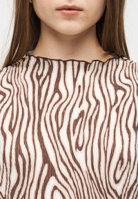 Gros plan sur le bas du visage et le cou d'une personne portant un haut plissé à rayures zébrées marron et blanc avec un col à volants.