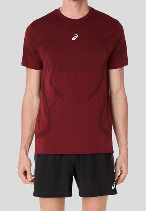 Homme portant un t-shirt de sport bordeaux à manches courtes et un short noir, tous deux avec un logo blanc Asics, debout devant un fond uni.