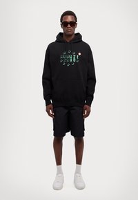 Black hoodie featuring green graphic text and star motifs, paired with black shorts and white sneakers. Model wearing sunglasses.