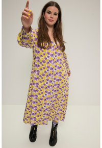 Vestido largo abotonado en blanco con un patrón floral en amarillo y morado. Presenta mangas largas y un ajuste relajado, combinado con botas negras hasta el tobillo.