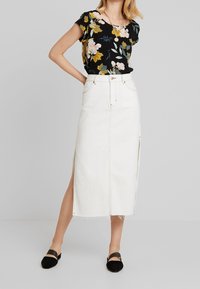Top nero floreale con maniche corte; gonna in denim bianco con spacchi laterali e cuciture a vista; abbinato a scarpe nere basse.
