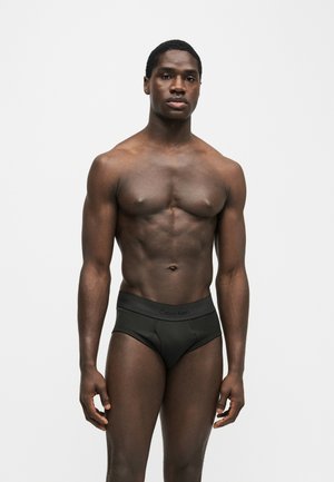 Homme musclé debout portant un slip Calvin Klein noir sur un fond uni gris clair, faisant face à l’avant avec une expression neutre.
