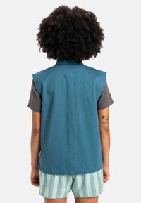 Tealfärgad ärmlös väst med hög krage, bärs över en grå t-shirt. Har en slät textur och platta sömmar. Randiga shorts syns nedanför.