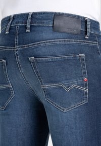 Jeans en denim bleu foncé avec une patch en cuir portant l'inscription "MAC JEANS" à la taille, avec des coutures blanches contrastantes et deux poches arrière.