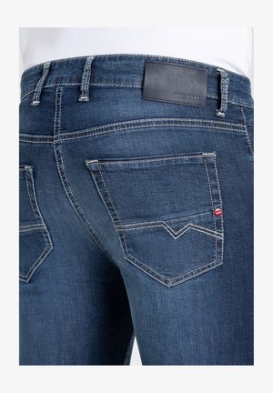 Jeans en denim bleu foncé avec une patch en cuir portant l'inscription "MAC JEANS" à la taille, avec des coutures blanches contrastantes et deux poches arrière.