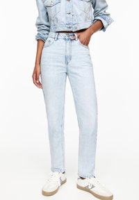 Jean en denim bleu clair à taille haute, coupe droite slim, avec poches avant et légère décoloration. Porté avec des baskets blanches.