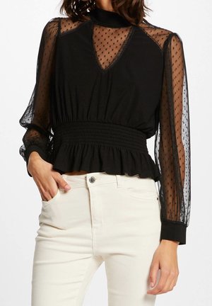 Schwarze transparente Bluse mit Pünktchenmuster, geraffter Taille und Rüschensaum, kombiniert mit hoch taillierter, cremefarbener Hose, die linke Hand in der Tasche.