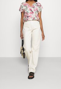 Kobieta idąca w krótkim rękawie kwiatowej bluzce, spodniach w kolorze kremowym z wysokim stanem, czarnych sandałach i niosąca małą torebkę o fakturze.