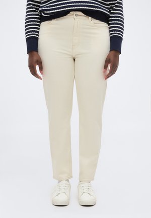 Person iført kremhvit tettsittende bukser, hvite joggesko og en marineblå og hvitstripet langermet topp, stående mot en ensfarget bakgrunn.