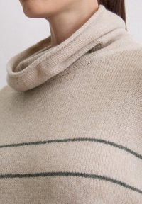 Beżowy sweter z dekoltem typu cowl, wykonany z miękkiego materiału, z dwiema ciemnozielonymi poziomymi paskami. Gładka struktura i luźny krój.