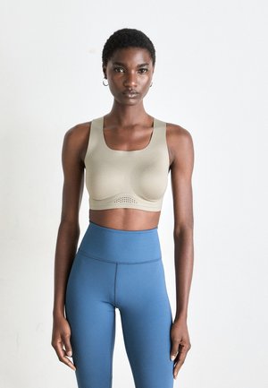 Kvinne med kort hår iført beige sportstrikk og høyt liv blå tights, stående mot en enkel lys bakgrunn.