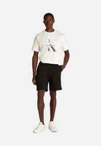 Maglietta bianca in cotone con stampa grafica e pantaloni corti neri. Indossati con sneakers bianche. Design semplice, vestibilità rilassata, senza motivi o accenti visibili.
