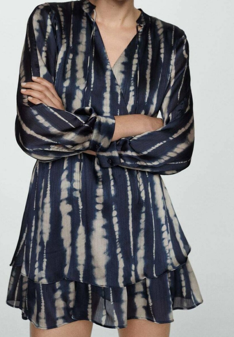 Femme portant une robe à manches longues tie-dye bleue et beige avec une jupe à couches, debout les bras croisés sur un fond uni.