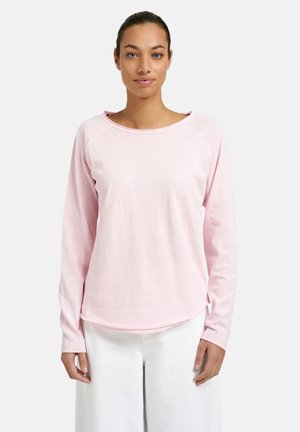 Woman wearing light pink long-sleeve shirt and white pants, standing against a plain light background, facing forward.