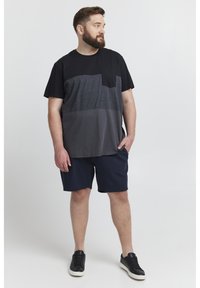 Schwarz-graues, farblich abgesetztes T-Shirt mit Brusttasche, kombiniert mit marineblauen Shorts. Das Modell trägt schwarze Sneakers. Weicher, lässiger Stoff.