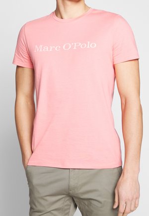Mężczyzna noszący różowy t-shirt z krótkim rękawem z tekstem "Marc O'Polo", w połączeniu z jasnoszarymi spodniami, ręce w kieszeniach, stojący na neutralnym tle.
