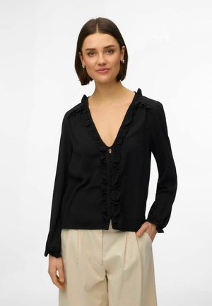 Kvinne med kort brunt hår iført en svart bluse med volanger og beige høyt liv med plisserte bukser, stående foran en hvit bakgrunn.