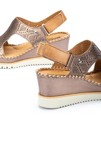 Sandalias cuña con cuero bronce perforado, correas en color marrón claro en el tobillo y una suela de goma blanca contrastante con un detalle de costura.
