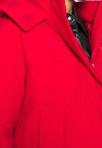 Czerwona pikowana kurtka z wysokim kołnierzem, z zapięciem na zamek błyskawiczny i kieszeniami na klapkę. Posiada gładką teksturę i kontrastowe czarne wykończenie.