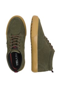 Zapatillas de ante verde con suelas de goma marrón, que presentan cordones redondos y detalles perforados en la puntera. Branding sutil visible en el interior.