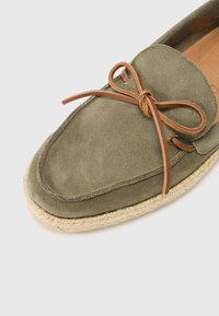 Scarpe in suede verde oliva con un fiocco in pelle color cuoio, forma rotonda e suola in corda in stile espadrillas. Texture liscia e dettagli a contrasto visibili.