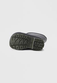 Suela de bota de goma negra con un patrón de textura, que presenta una entresuela en un tono gris verdoso y el logo de la marca embosado en el lateral.