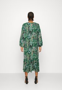 Grön klänning med paisleymönster, långa ärmar, rynkad fåll och lös passform, tillverkad av ett lätt tyg. Svarta klacksandaler bärs.