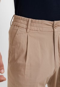 Beżowe bawełniane spodnie z elastycznym paskiem w talii, plisami z przodu oraz zapinane na pojedynczy guzik. Gładka faktura, luźny krój, casualowy styl.