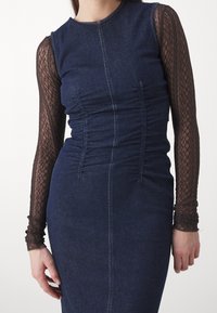 Džínové šaty s řaseným předním dílem, tmavě modré látky a přiléhavého střihu; mají dlouhé černé krajkové rukávy s texturovaným vzorem.