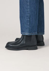 Bottes noires en cuir avec panneaux latéraux élastiques et semelle en caoutchouc texturée, associées à un jean bleu denim raccourci.