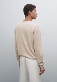 Maglione beige a maniche lunghe con scollo rotondo e polsini a coste, indossato con pantaloni bianchi. Texture liscia, vestibilità rilassata e design minimal.