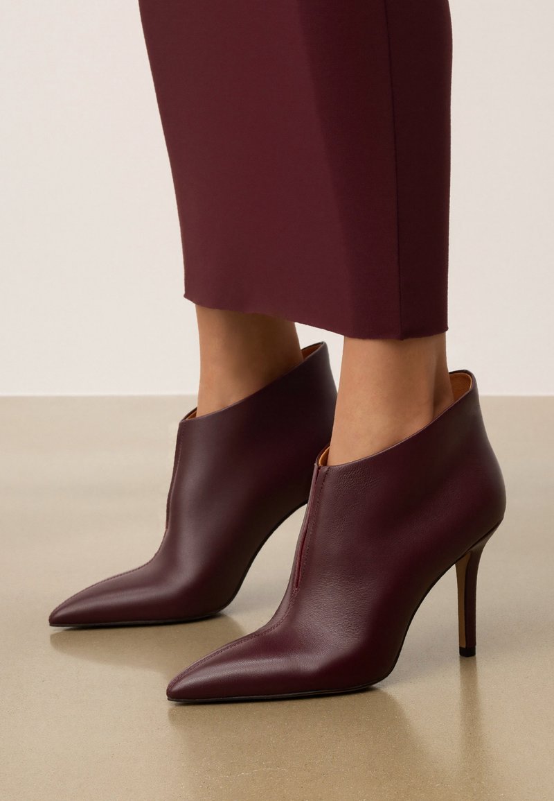 Botas de tobillo de cuero burdeos con punta afilada, cremallera lateral y tacón delgado. Textura suave y diseño contemporáneo.