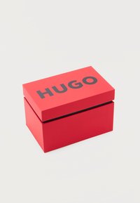 Κόκκινο ορθογώνιο κουτί με μαύρο κείμενο "HUGO" στο καπάκι, τοποθετημένο σε απλό λευκό φόντο.