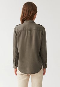 Camisa de manga larga verde oliva con cuello tipo polo, puños abotonados y dobladillo curvado. Tela suave con un ajuste ligeramente holgado y hombreras.