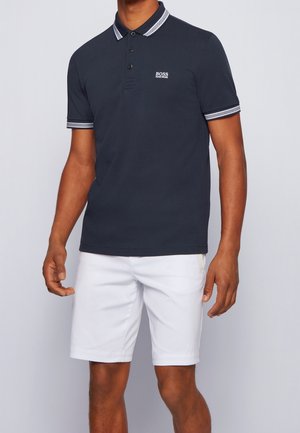 Homme portant un polo bleu marine avec col et manches rayés blancs, assorti à un short blanc mi-genou.