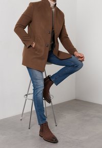 Brun ullblandad kappa med hög krage, två knappar och sidofickor, i kombination med ljusblå jeans och bruna anklarstövlar, sittande på en pall.